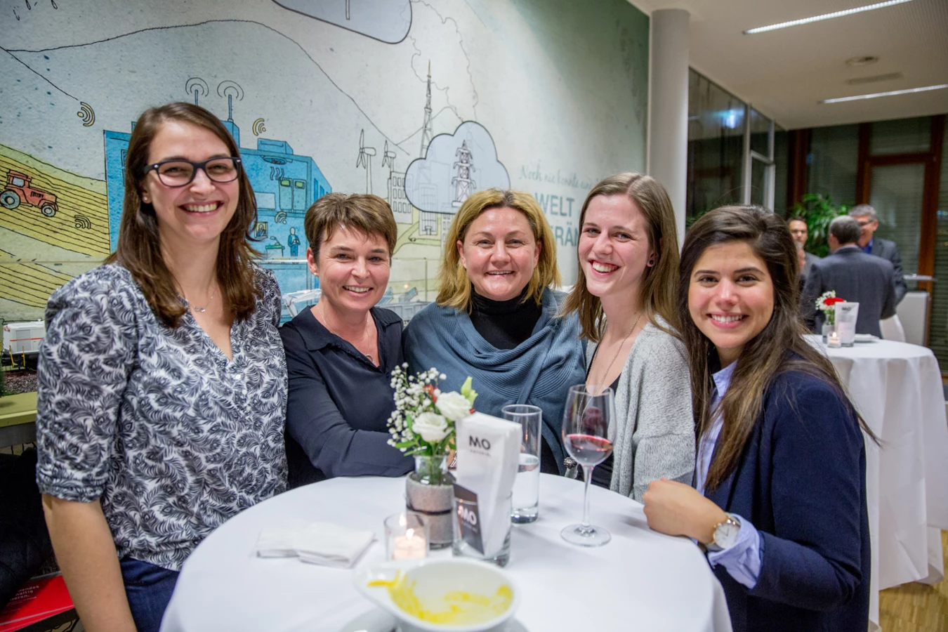 Fünf Frauen sitzen lachend an einem Stehtisch mit Getränken bei einer Veranstaltung, mit Wandgemälde im Hintergrund (Hubert, Geburtstag).