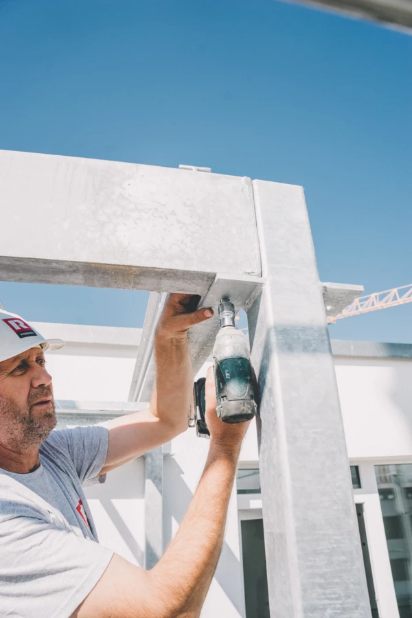 Ein Bauarbeiter in einem weißen Helm mit rotem Rhomberg-Logo schraubt ein Metallteil an einer Baustelle fest, während die Sonne scheint (Hochbau).