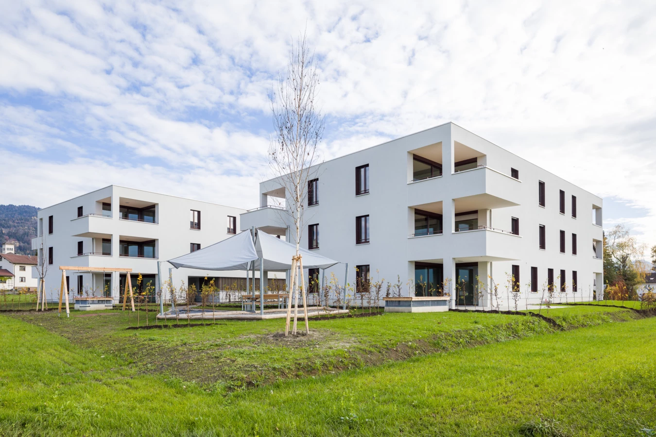 Zwei moderne weiße Wohngebäude mit Balkonen, umgeben von grünen Rasenflächen, mit einem überdachten Sitzbereich und jungen Bäumen (Unterhochstegstrasse).