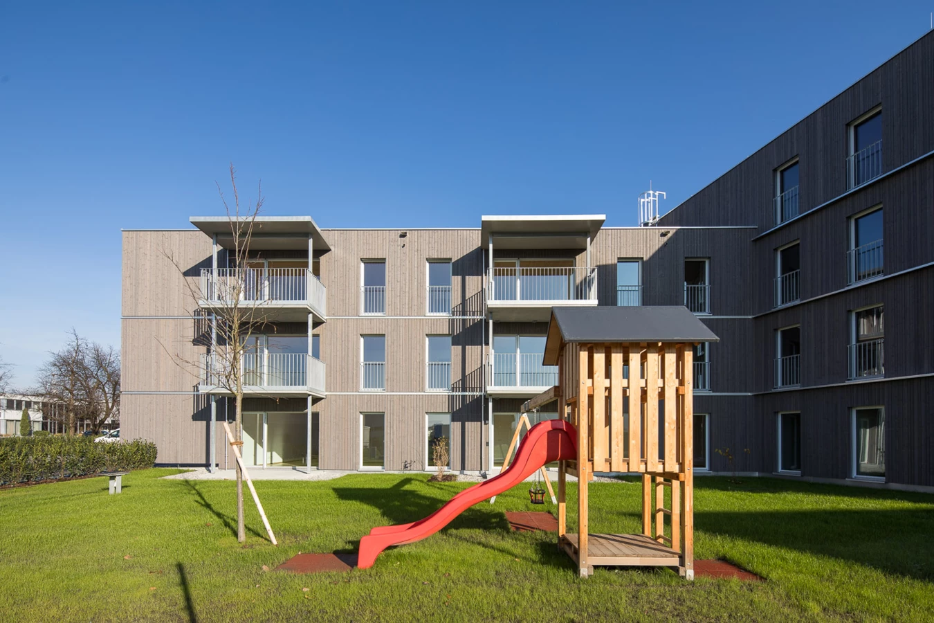 Modernes Mehrfamilienhaus mit Holzfassade, Balkonen und einem Spielplatz mit roter Rutsche bei sonnigem Wetter (Froschweg).