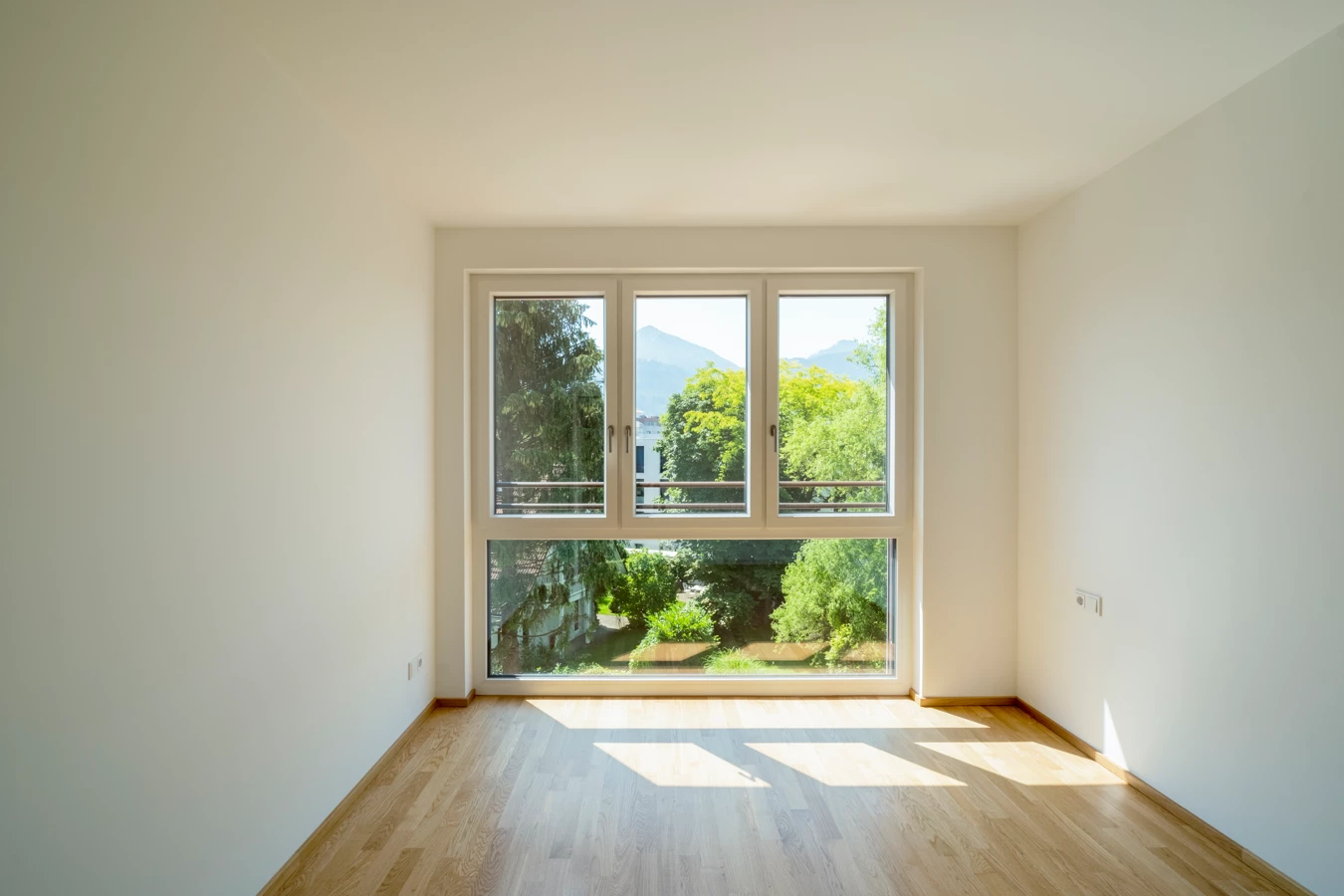 Leeres Zimmer mit Holzboden und einem großen Fenster, das auf grüne Bäume und Berge in Dornbirn blickt