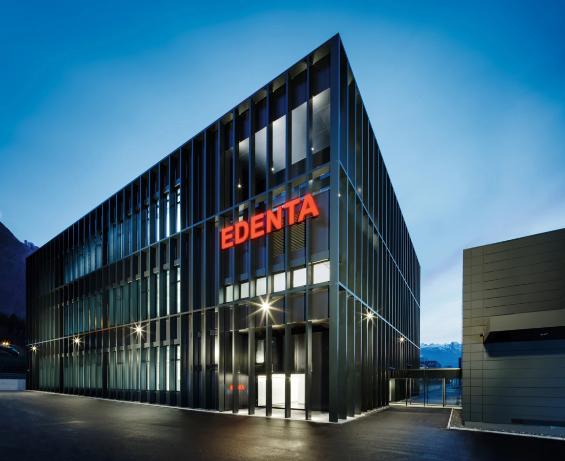 Modernes Geschäftsgebäude von Edenta mit horizontalen und vertikalen Lamellen, beleuchtet in der Dämmerung unter blauem Himmel.