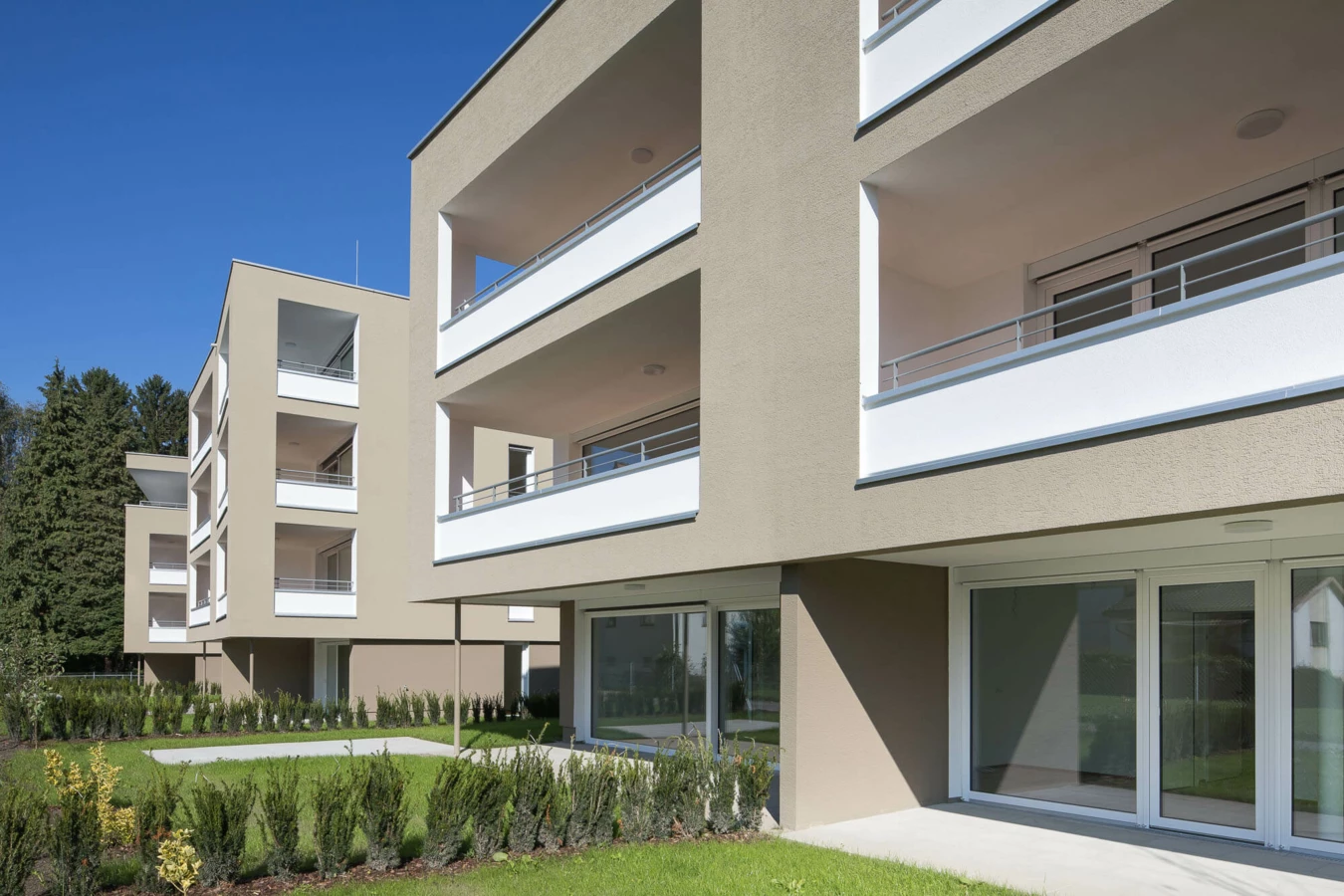 Moderne, hellbraune Wohngebäude mit weißen Balkonen und großen Fenstern, umgeben von grünen Rasenflächen und Hecken unter blauem Himmel in der Wohnanlage Widum Lustenau.