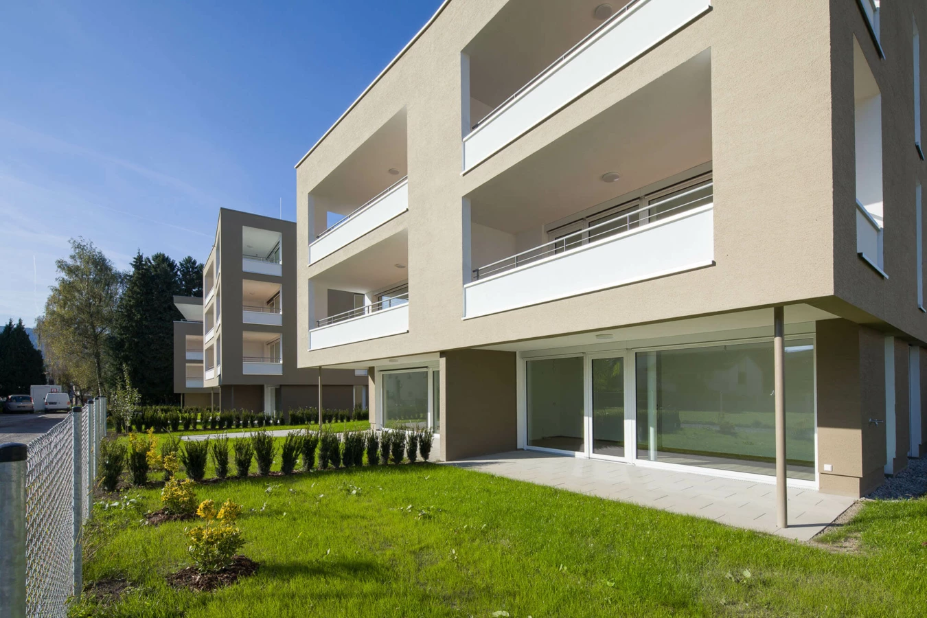 Moderne Wohnanlage Widum in Lustenau mit mehreren Gebäuden, gepflegten Rasenflächen und Hecken unter blauem Himmel.