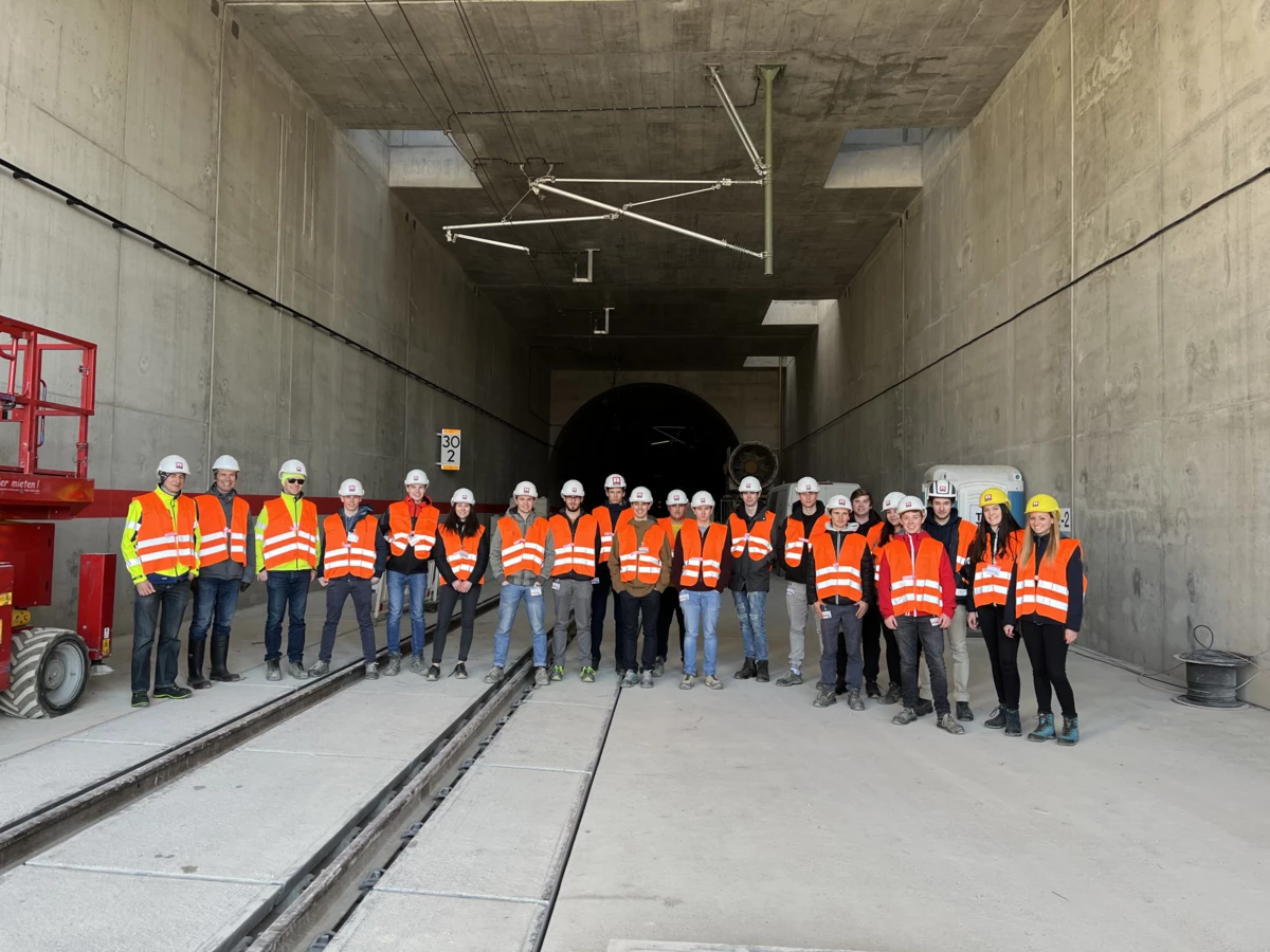 Eine Gruppe von 18 Personen in Sicherheitskleidung mit Helmen und Warnwesten steht vor einem Tunnelportal mit Gleisen (Rankweil, Deutschland, Gruppenfoto).