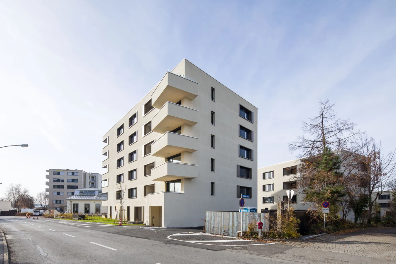 Modernes, mehrstöckiges Wohngebäude mit Balkonen an einer Straße, flankiert von Bäumen und weiteren Gebäuden unter blauem Himmel (Bäumleareal).