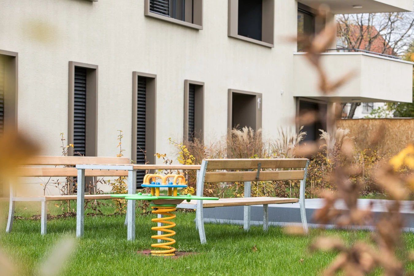 Spielplatz mit gelbem Wipptier und Holzbänken vor einem modernen Wohngebäude mit Schiebefenstern (Baeumlequartier)