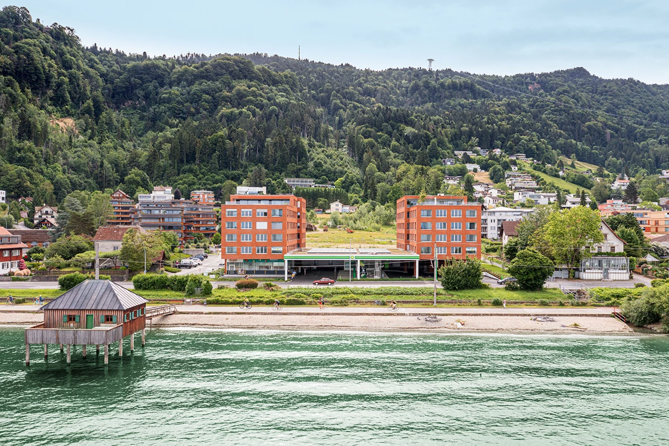 Zwei moderne rote Backsteingebäude mit einer Tankstelle dazwischen, am Ufer eines Sees mit Bergen und Bäumen im Hintergrund (Seeblick, Lochau).