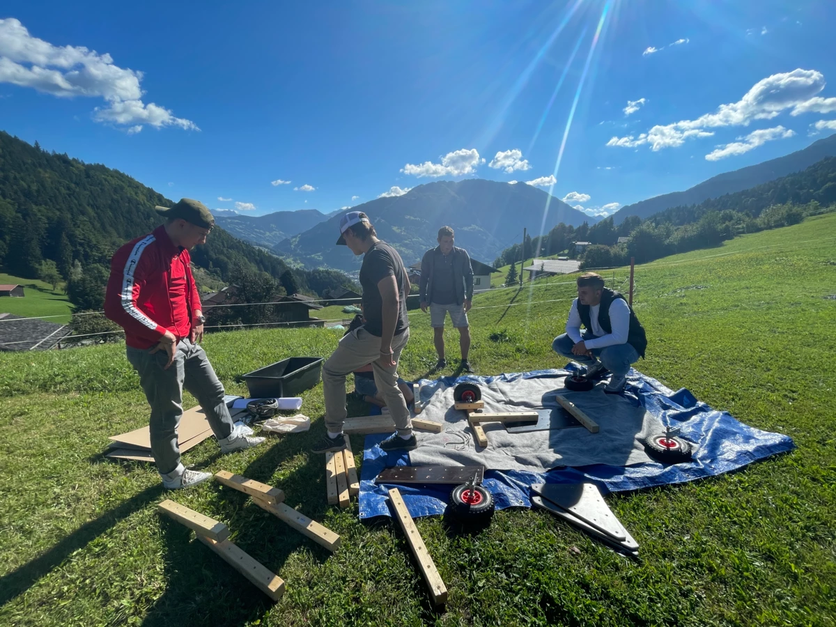 Vier junge Männer bauen im Freien auf einer blauen Plane ein Objekt zusammen, umgeben von grünen Wiesen und Bergen unter blauem Himmel mit Sonnenschein (Lehre).