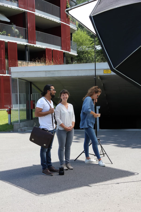 Zwei Frauen und ein Mann posieren lächelnd für ein Fotoshooting vor einem Gebäude mit roten Fassaden und einer Garageneinfahrt.