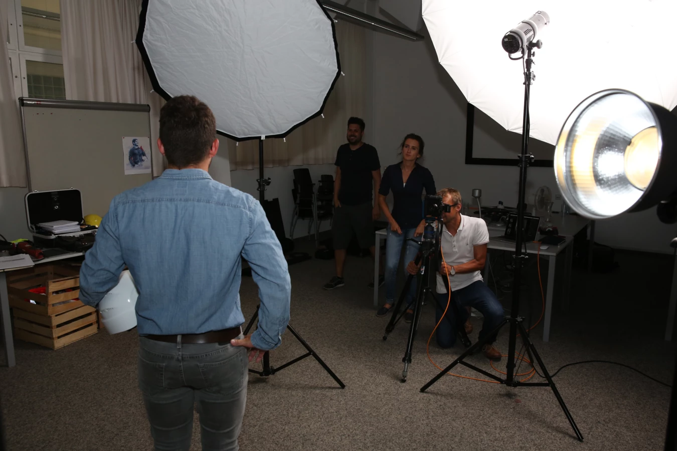 Ein Fotoshooting-Set mit einem Mann, der eine Bauarbeiter-Sicherheitskappe hat, vor Softboxen, während ein Kameramann filmt.