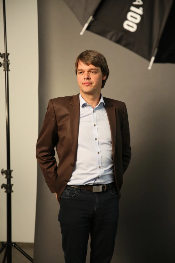 Ein Mann im braunen Sakko und hellblauem Hemd posiert vor einem dunkelgrauen Fotohintergrund.