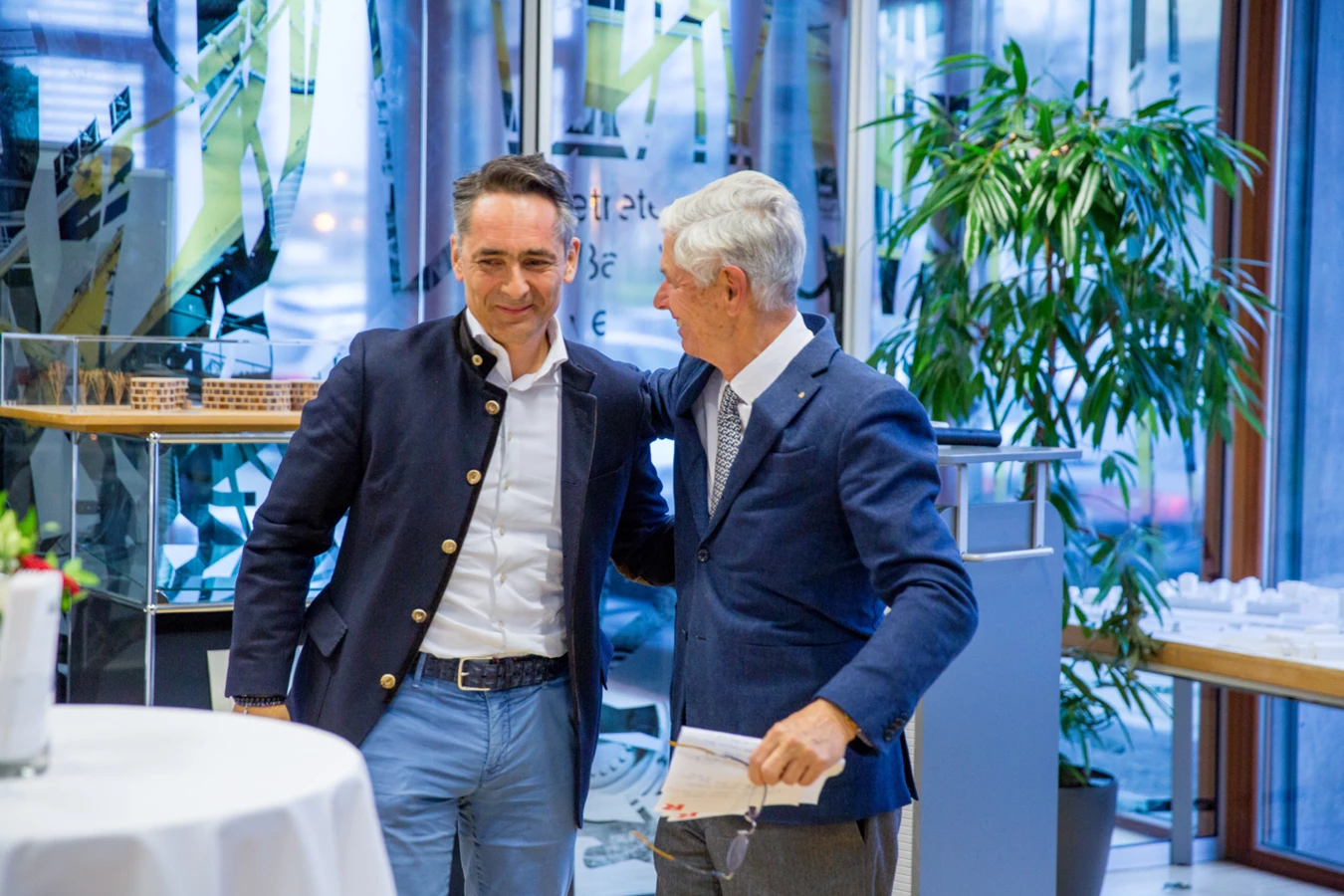 Zwei Männer in Anzügen unterhalten sich lachend bei einer Veranstaltung, mit Pflanzen und Architekturmodellen im Hintergrund (Hubert, Geburtstag).