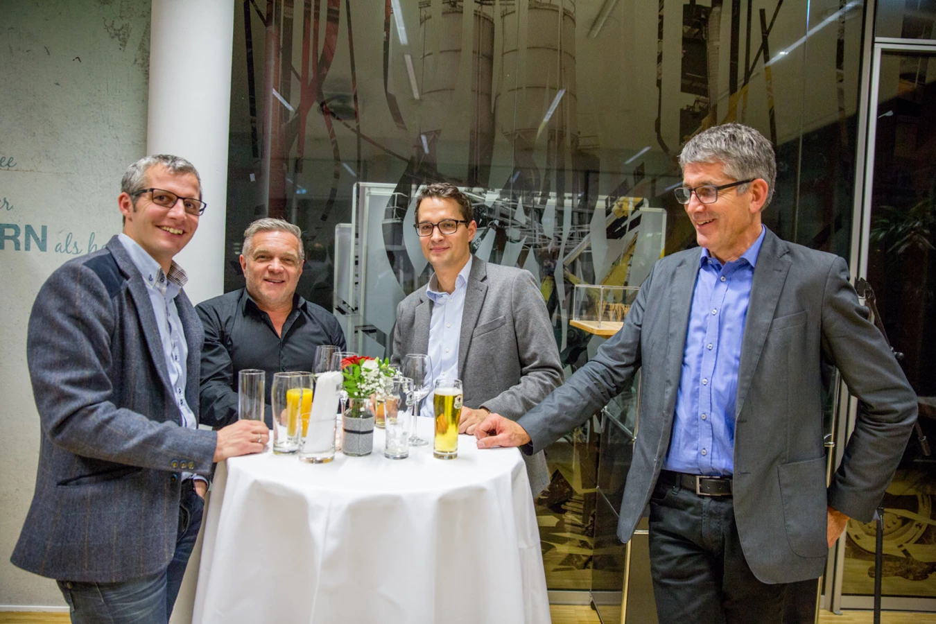 Vier Männer in Anzügen stehen lachend um einen Stehtisch mit Getränken bei einer Veranstaltung (Hubert, Geburtstag).