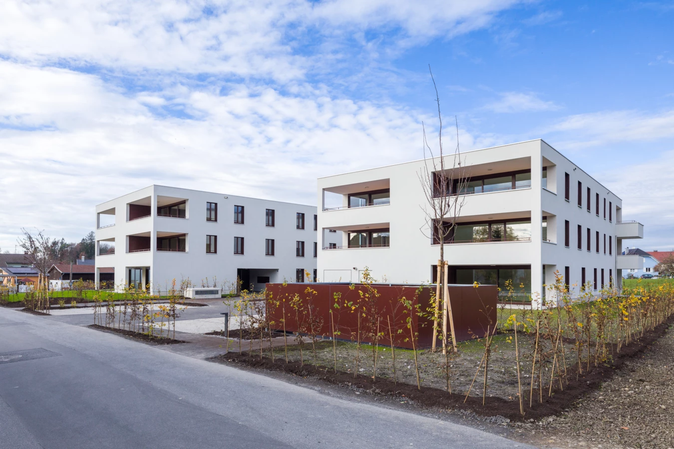 Zwei moderne, weiße Mehrfamilienhäuser mit Balkonen und Fenstern, umgeben von jung gepflanzten Bäumen und Sträuchern an der Unterhochstegstraße.