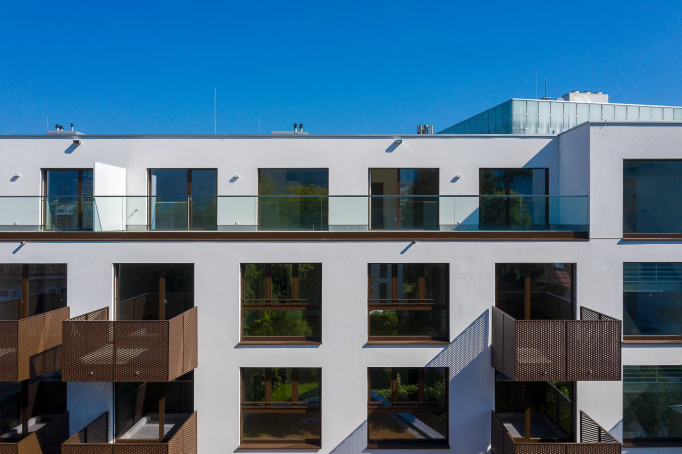 Fassade eines weißen Gebäudes mit braunen perforierten Metallbalkonen und großen Fenstern unter blauem Himmel (Dornbirn).