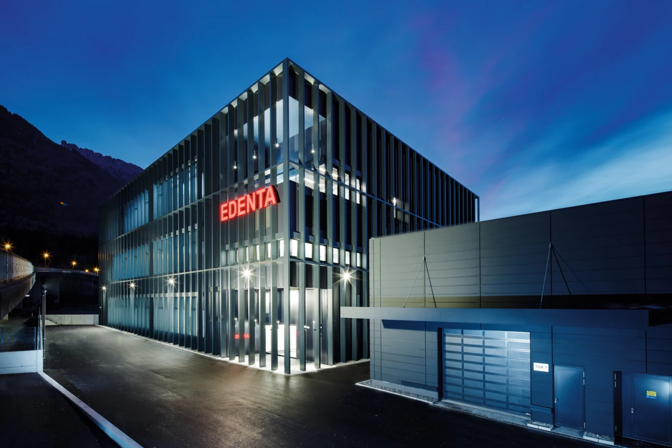 Modernes Geschäftsgebäude von Edenta bei Dämmerung mit beleuchtetem Logo und klarer Architektur unter blauem Himmel.