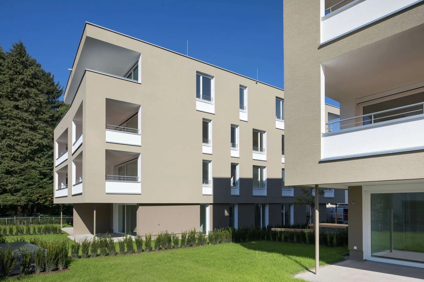 Zwei moderne helle Wohnanlagen Widum Lustenau auf einer grünen Wiese mit Bäumen unter blauem Himmel.