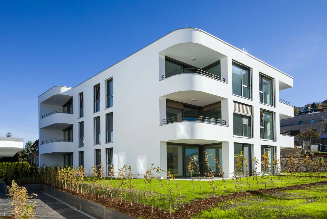 Moderne weiße Wohnanlage mit abgerundeten Balkonen, großen Fenstern, grünen Rasenflächen und jungen Bäumen unter blauem Himmel in Feldkirch (Wolf-Huber-Straße).