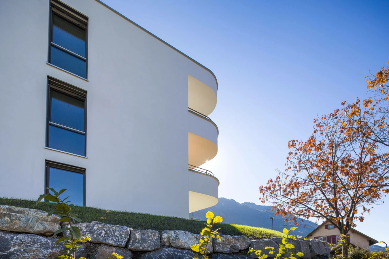 Modernes weißes Gebäude mit runden Balkonen und Fenstern über einer Steinmauer, Herbstbaum und Berge im Hintergrund unter blauem Himmel in Feldkirch (Wolf-Huber-Straße, Wohnanlage).