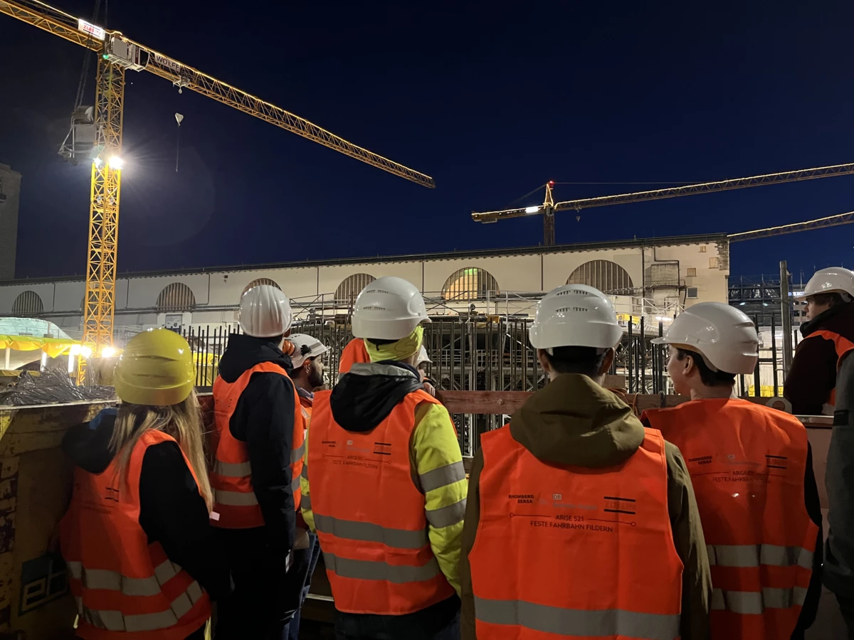 Arbeiter in Warnwesten und Helmen betrachten bei Nacht eine Baustelle mit Kranen und Gebäuden in Fildern (Rankweil, Deutschland, Kran).