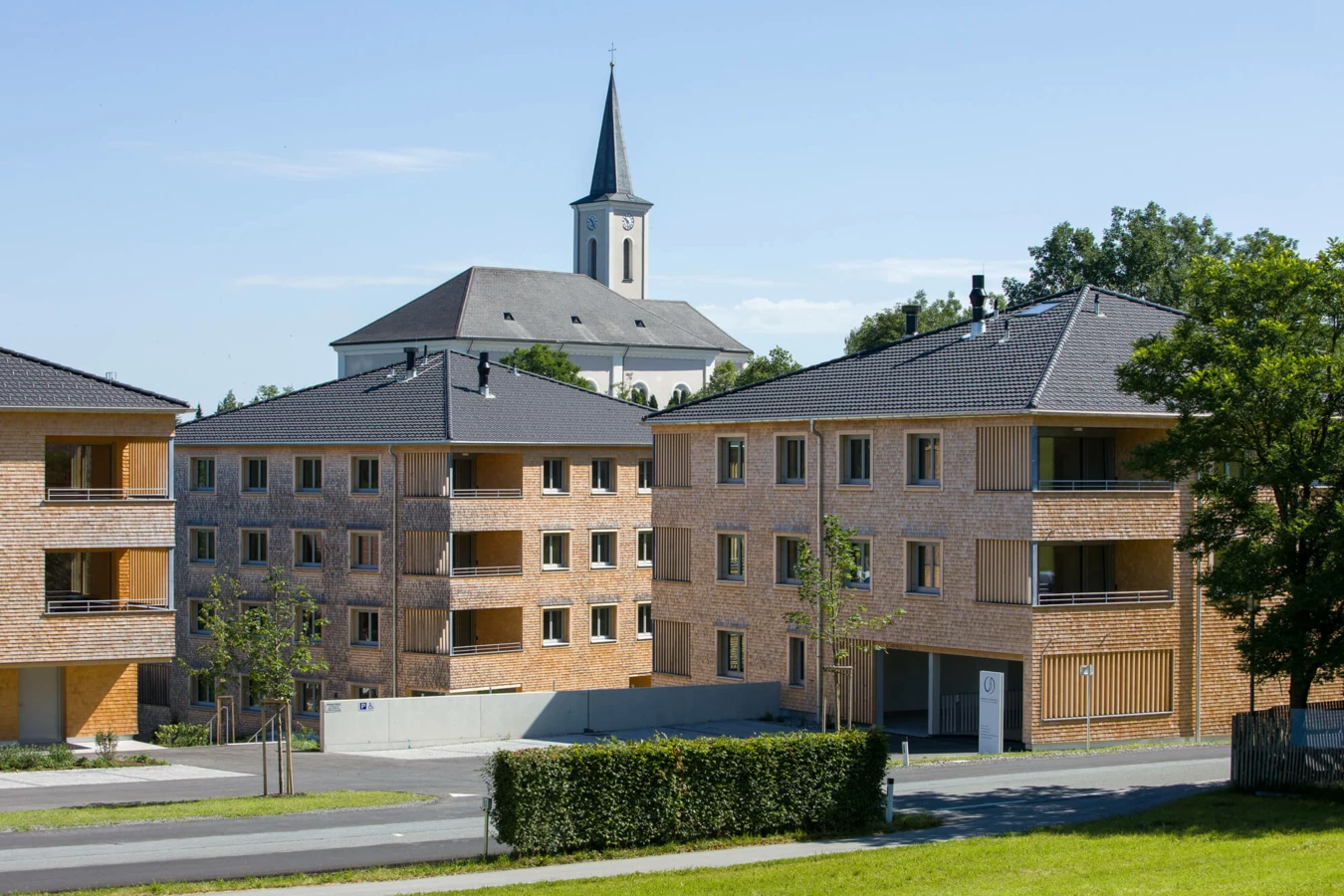 Zwei moderne Mehrfamilienhäuser mit Holzfassade und grauen Dächern in Alberschwende, mit Kirche im Hintergrund (Wohnanlage).