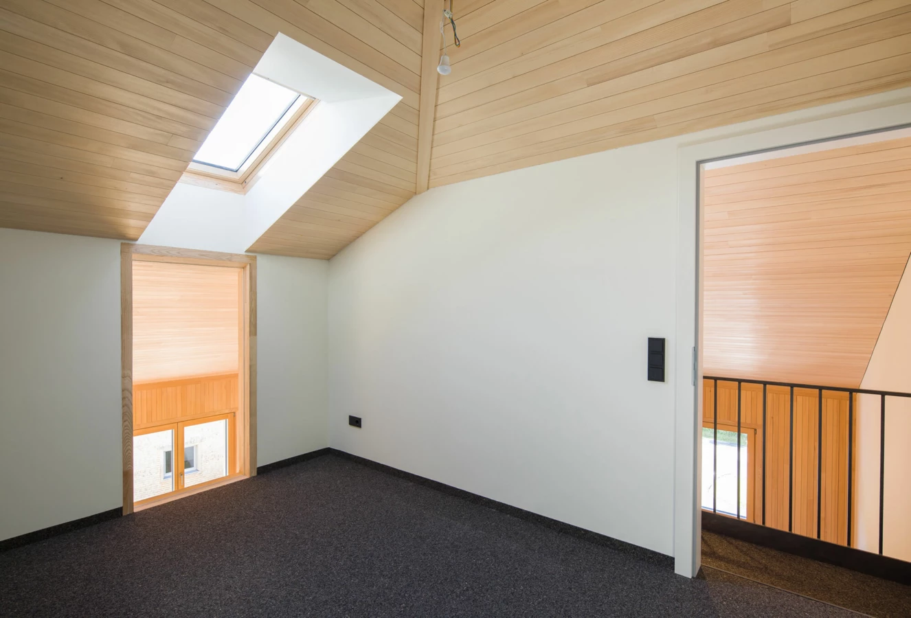 Dachgeschosszimmer mit Holzbalkendecke, schrägem Dachfenster, modernem Teppichboden und Fenster zum Treppenhaus. Eine Glühbirne hängt an der Decke (Wohnanlage, Alberschwende).