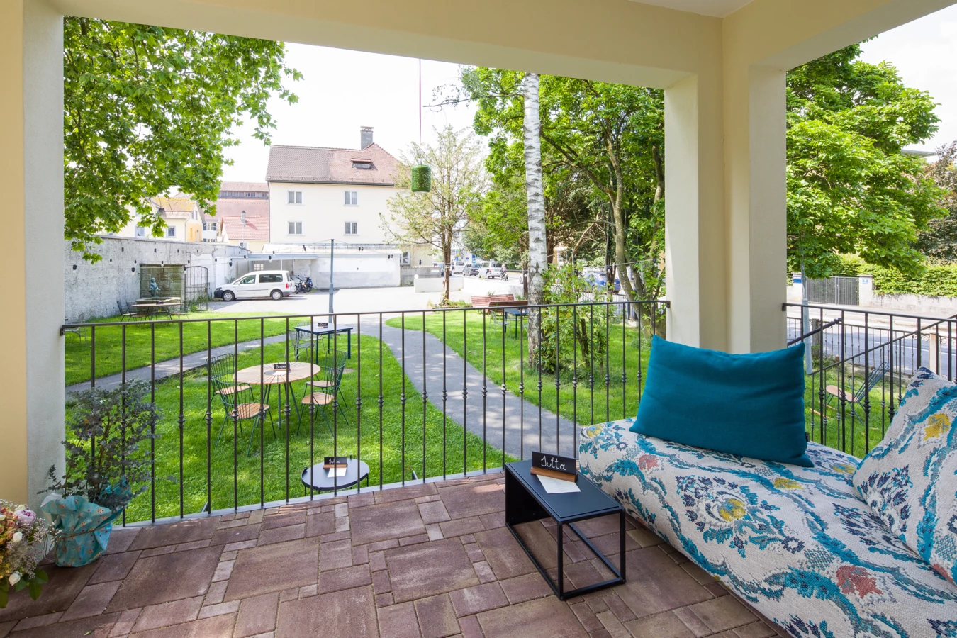 Blick von einer Terrasse mit gemusterter Liege und türkisfarbenem Kissen auf einen begrünten Garten mit Café-Tischen und einem Weg (Wunderkind, Backstube).