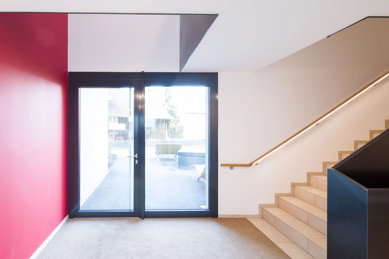 Eingangsbereich mit Glastür zu einem Außenbereich, einer roten Wand, Treppen und einem beleuchteten Holzgeländer (Bäumleareal).