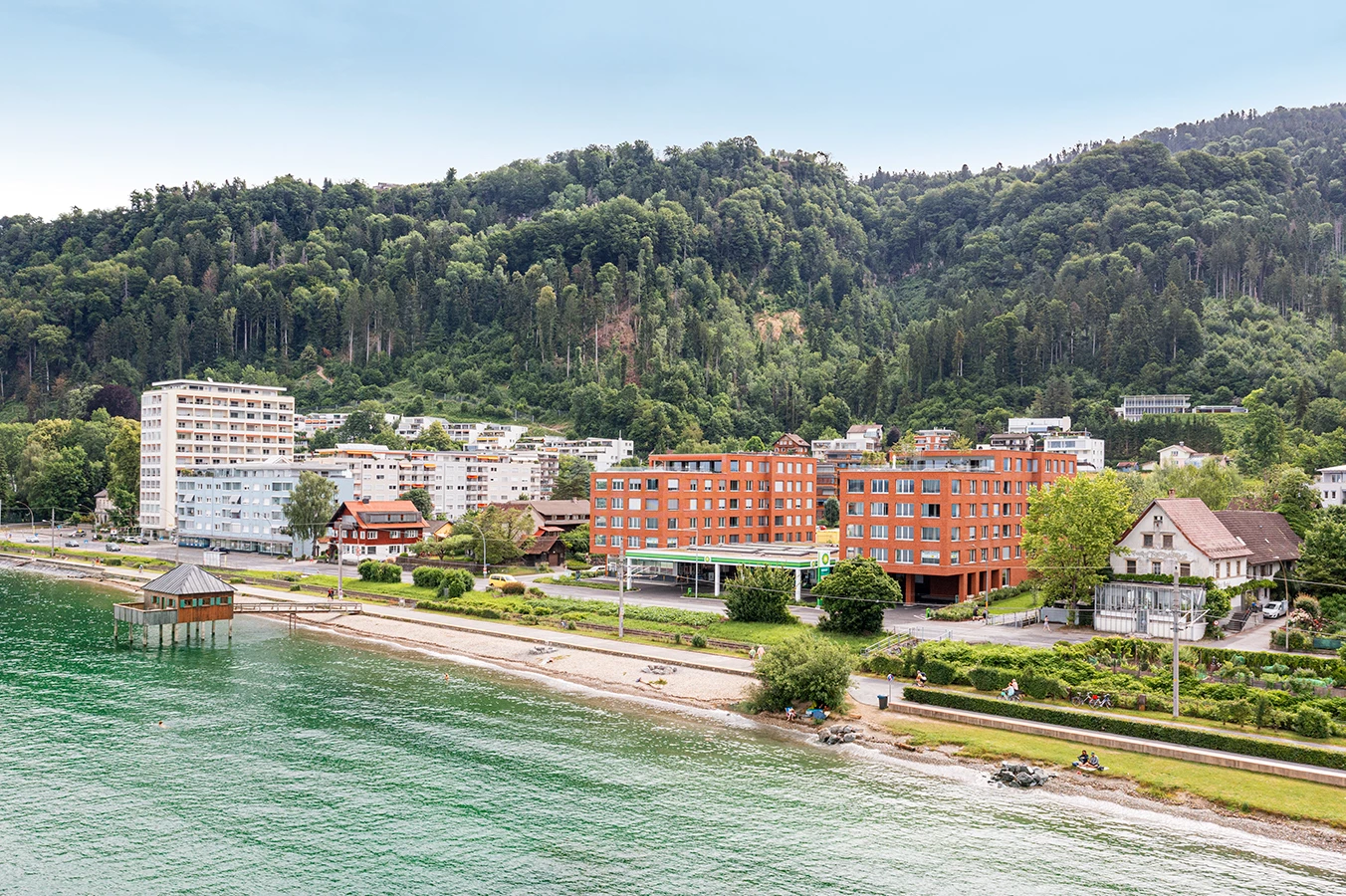 Mehrere Wohngebäude am Ufer des Bodensees in Lochau, darunter neue rote Gebäude und ältere weiße Türme, mit bewaldeten Hügeln im Hintergrund (Seeblick).