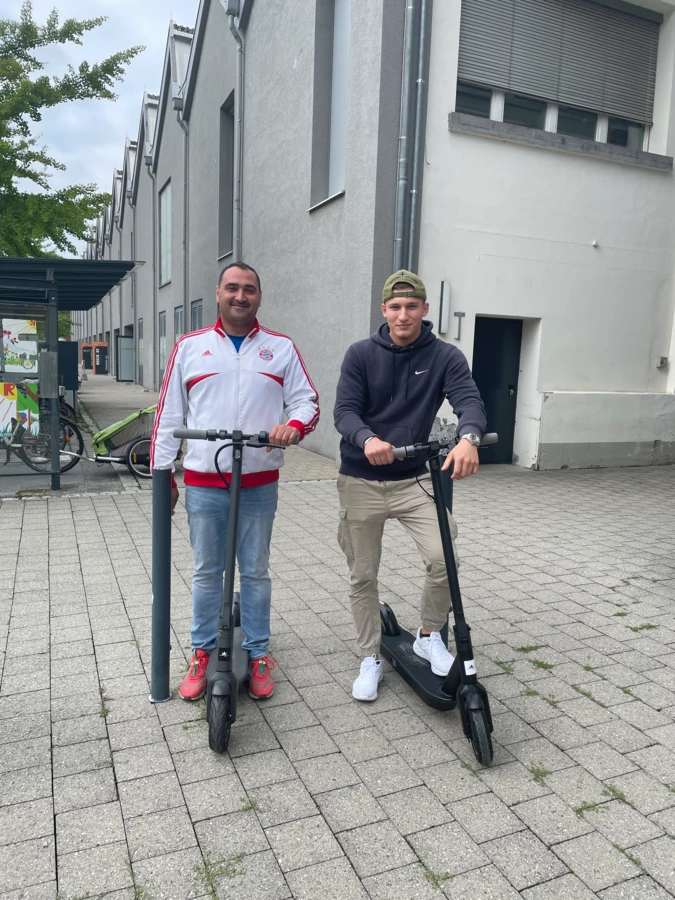 Zwei junge Männer stehen lächelnd mit schwarzen E-Scootern auf einem gepflasterten Gehweg vor modernen Gebäuden, links ein Fahrradständer (Lehre).