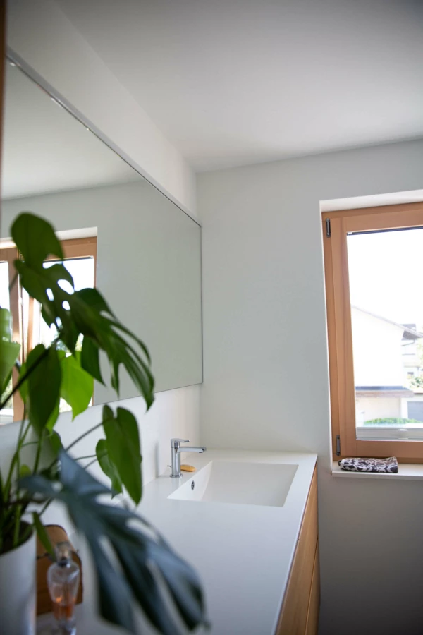 Modernes Badezimmer mit Waschtisch, großem Spiegel, Holzfenster und einer Grünpflanze. Das neue Bad in Dornbirn (Generalsanierung, Einfamilienhaus).