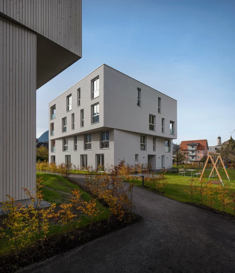 Moderne Wohnanlage Zürcherstrasse Bludenz mit weiß verputzten Gebäuden und Holzverkleidung, umgeben von Grünflächen und einem Spielplatz an einem sonnigen Tag.