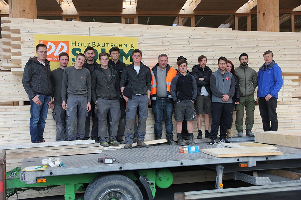 Eine Gruppe junger Männer und zwei ältere Männer posieren vor einem Stapel Holzbretter und einem Schild mit der Aufschrift "Holz Bautechnik Solm" (Lehrlingstag).