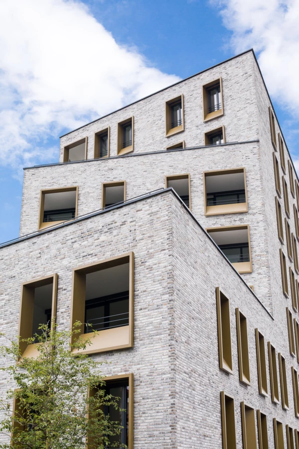 Modernes Gebäude in Dornbirn, Kreuzgasse 4, mit heller Ziegelfassade, eckigen, goldfarbenen Fensterrahmen und einem Baum im Vordergrund.