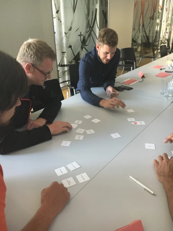 Drei Männer sitzen an einem Tisch und verschieben kleine Kärtchen, die mit Text bedruckt sind, in einem Workshop.