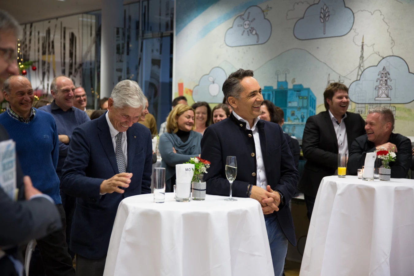 Eine Gruppe lachender, festlich gekleideter Männer und Frauen steht in einem Raum an Stehtischen mit Getränken (Hubert, Geburtstag).