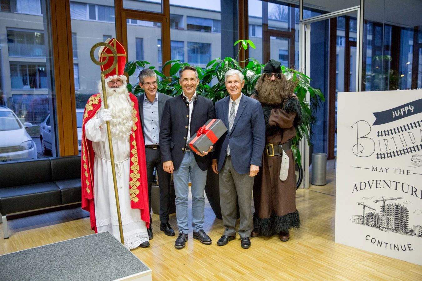 Vier Männer in Anzügen und Kostümen, darunter St. Nikolaus, stehen vor einer Zimmerpflanze und einem Geburtstagsplakat mit Bauvisualisierung (Hubert).