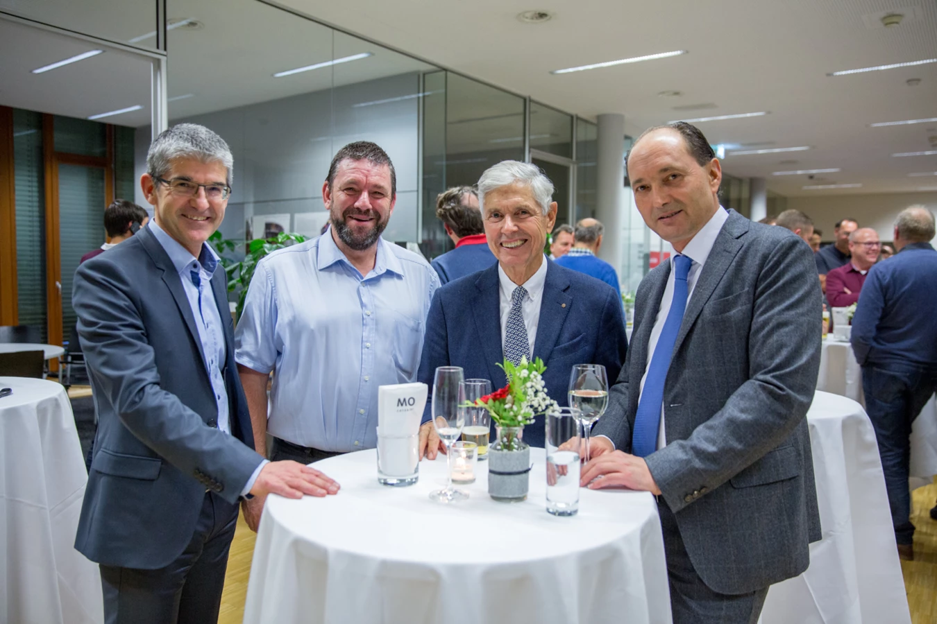 Vier lächelnde Männer in stilvoller Kleidung, die bei einer Veranstaltung an einem Stehtisch mit Getränken und Blumen stehen (Hubert, Geburtstag).