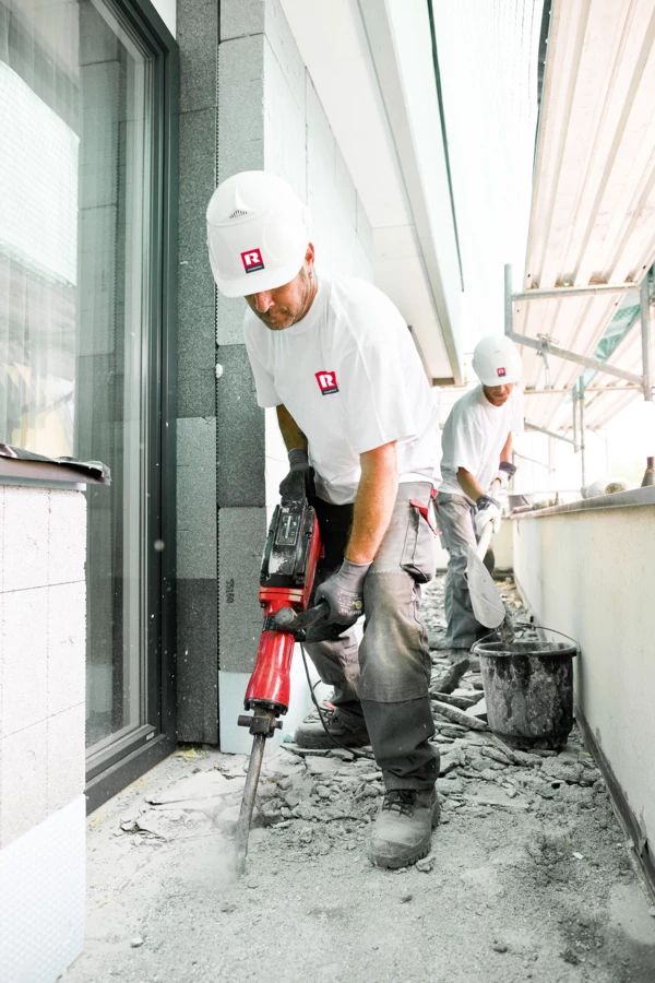 Zwei Bauarbeiter in Schutzkleidung auf einer Baustelle. Einer verwendet einen Bohrhammer, der andere schaufelt Schutt in einen Eimer (Hochbau).