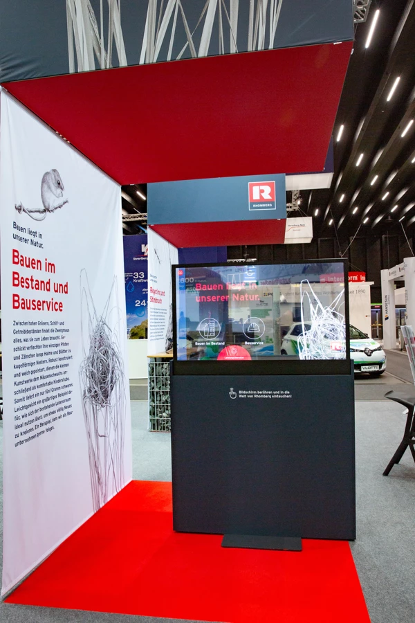 Rhomberg Bau Messestand mit interaktivem Bildschirm, rotem Teppich und Banner mit Mausmotiv und Text "Bauen liegt in unserer Natur"