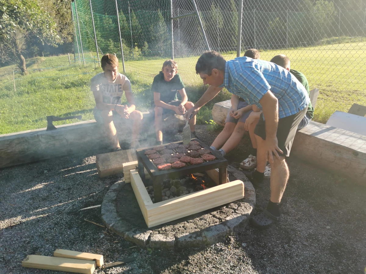 Ein Mann im blau karierten Hemd grillt Burger für eine Gruppe sitzender Jugendlicher beim Lehrlingscamp 2019.