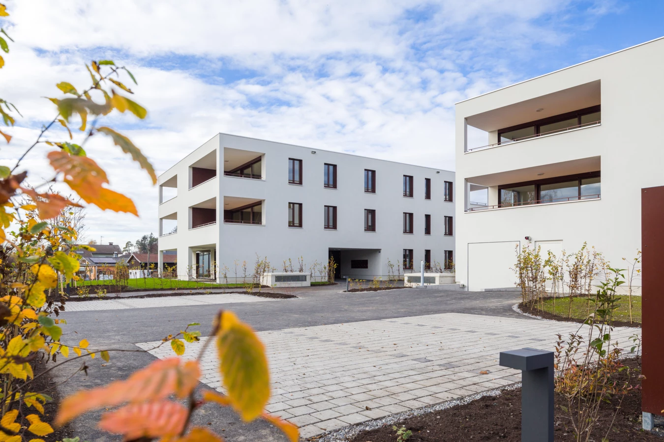 Zwei moderne weiße Mehrfamilienhäuser mit Balkonen und dunklen Fenstern, umgeben von gepflasterten Flächen und junger Bepflanzung unter blauem Himmel mit Wolken (Unterhochstegstrasse).