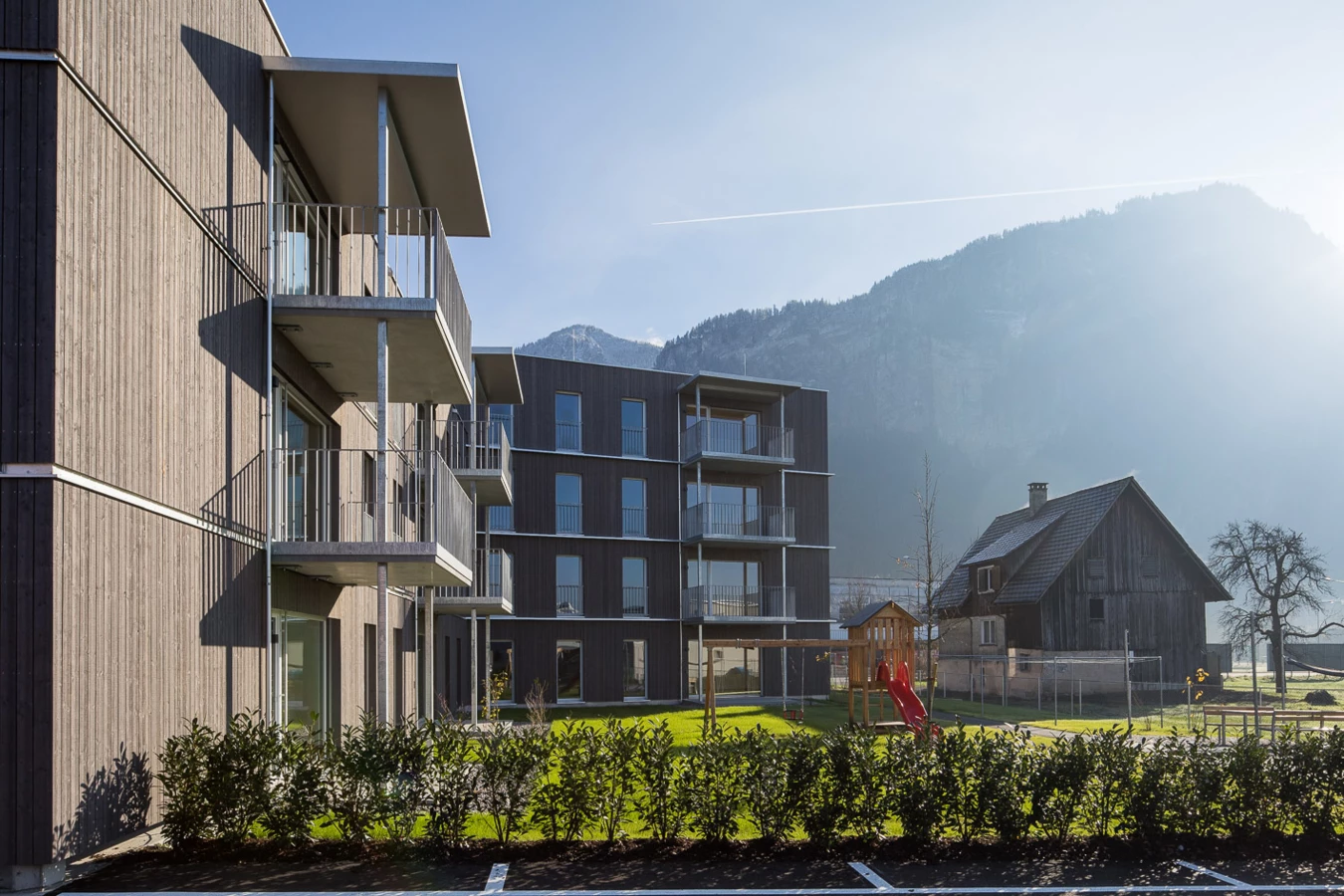 Moderne Wohngebäude mit Holzfassaden und Balkonen neben einem Spielplatz, einer Scheune und Bergen bei Sonnenlicht (Froschweg).