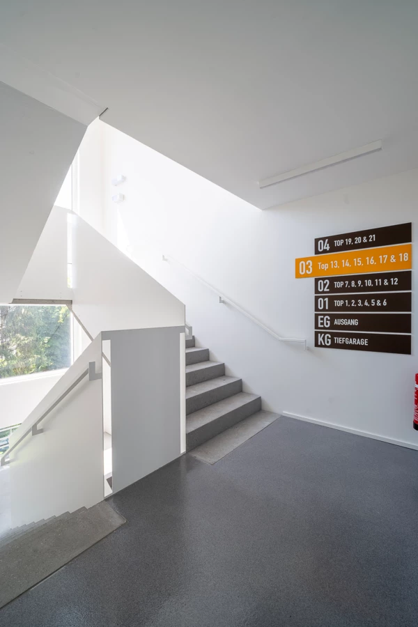 Modernes Treppenhaus in einem Gebäude mit dunkelgrauem Boden, weißen Wänden und einer Anzeigetafel für Stockwerke mit "EG Ausgang" und "KG Tiefgarage" (Dornbirn).