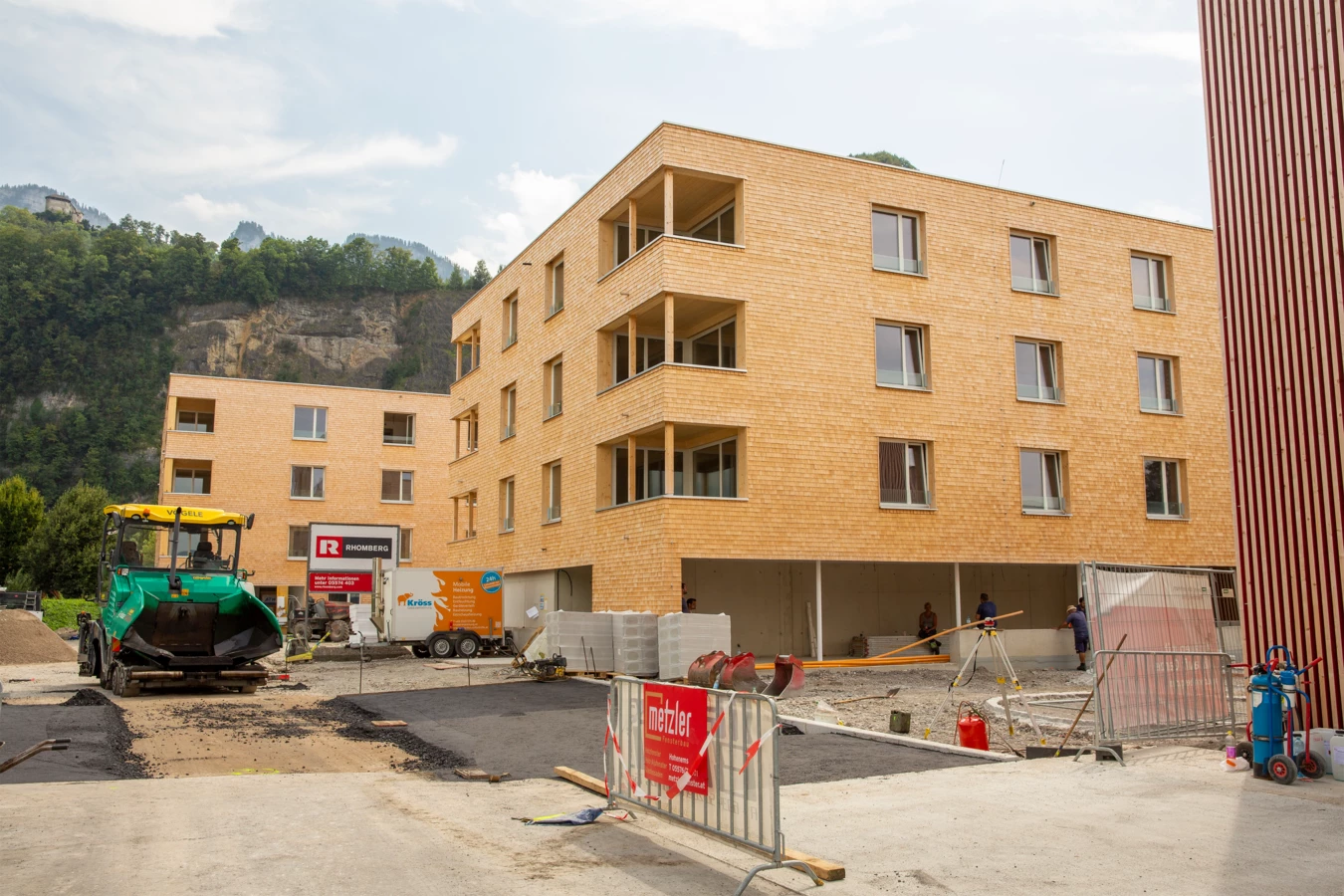 Zwei moderne Holzgebäude auf einer Baustelle mit einem grünen Asphaltfertiger und Bauarbeitern (Hellbrunnenstraße).