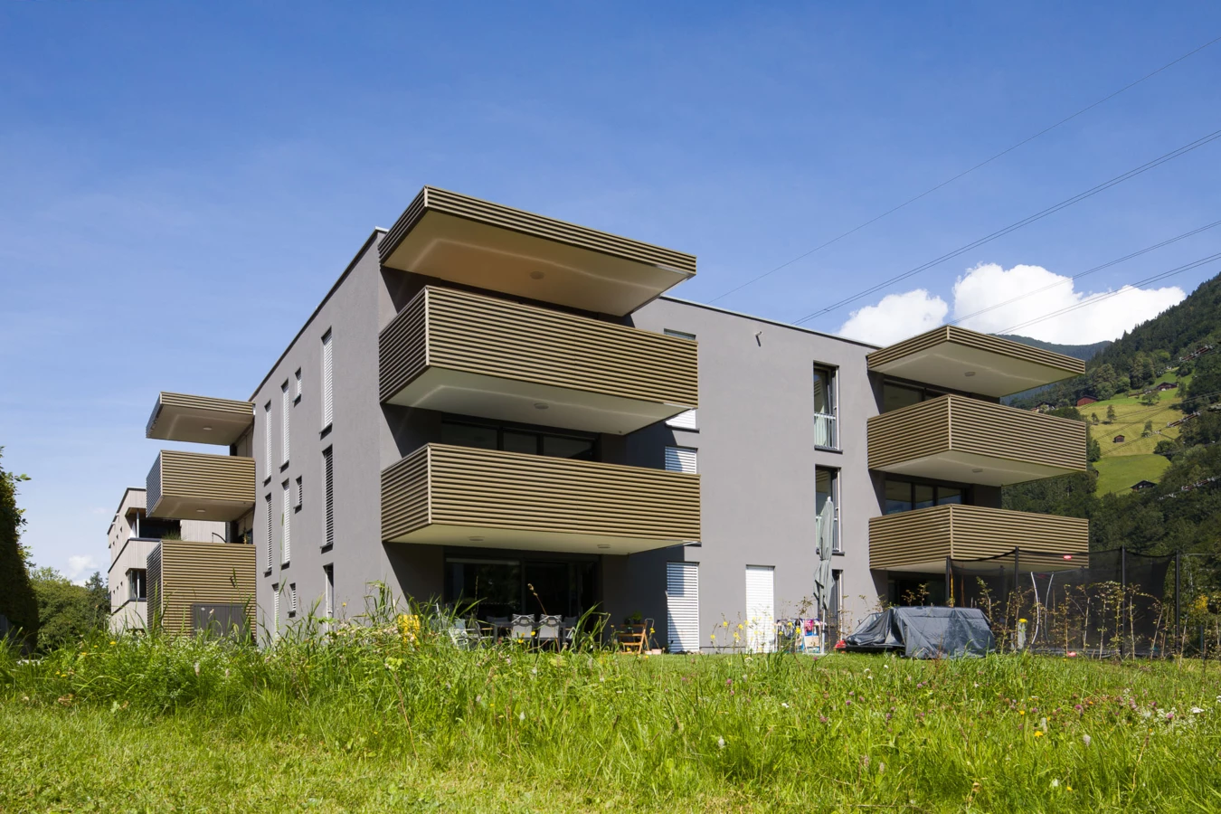 Moderne Wohnanlage in Vandans mit grauer Fassade, mehreren Balkonen mit Holzlamellen und einer grünen Wiese im Vordergrund unter blauem Himmel (Bahnhofstraße, Bahnhofsstrasse).