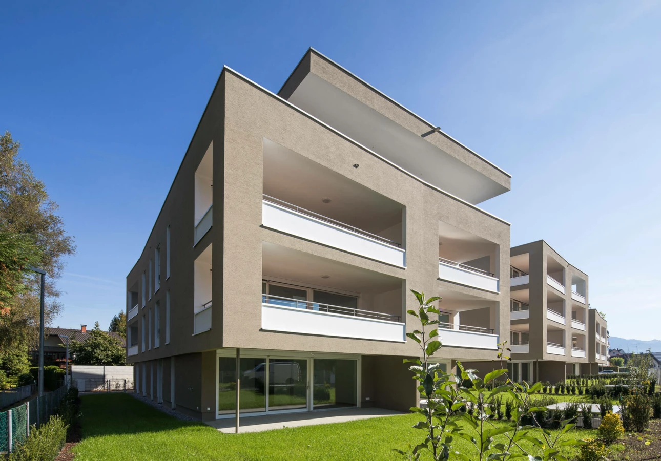 Moderne Wohngebäude der Wohnanlage Widum in Lustenau mit brauner Fassade, weißen Balkonen und gepflegtem Rasen unter blauem Himmel.