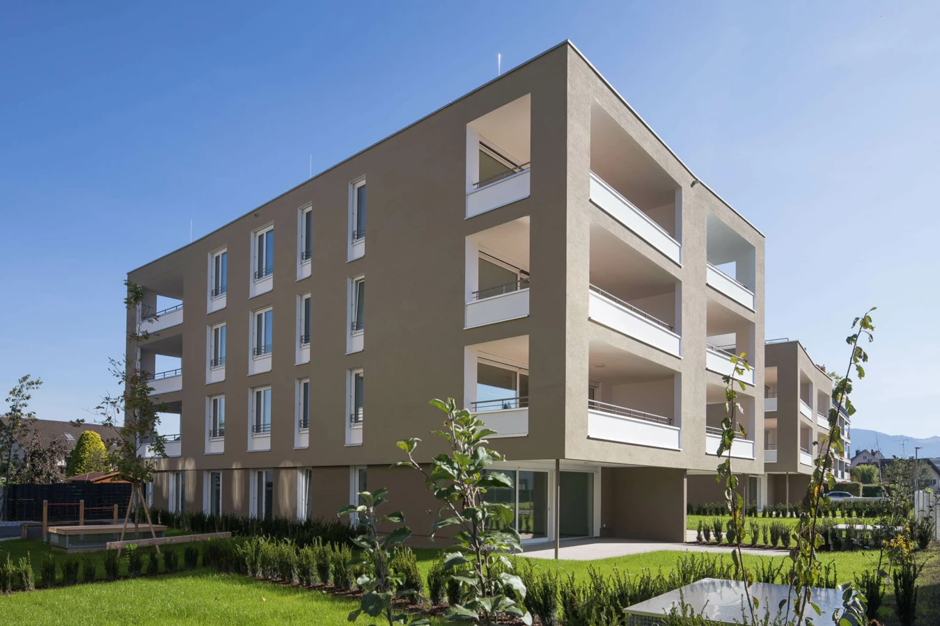 Moderne Wohnanlage Widum in Lustenau mit beigefarbenen Fassaden, weißen Balkonen und Grünflächen unter blauem Himmel.