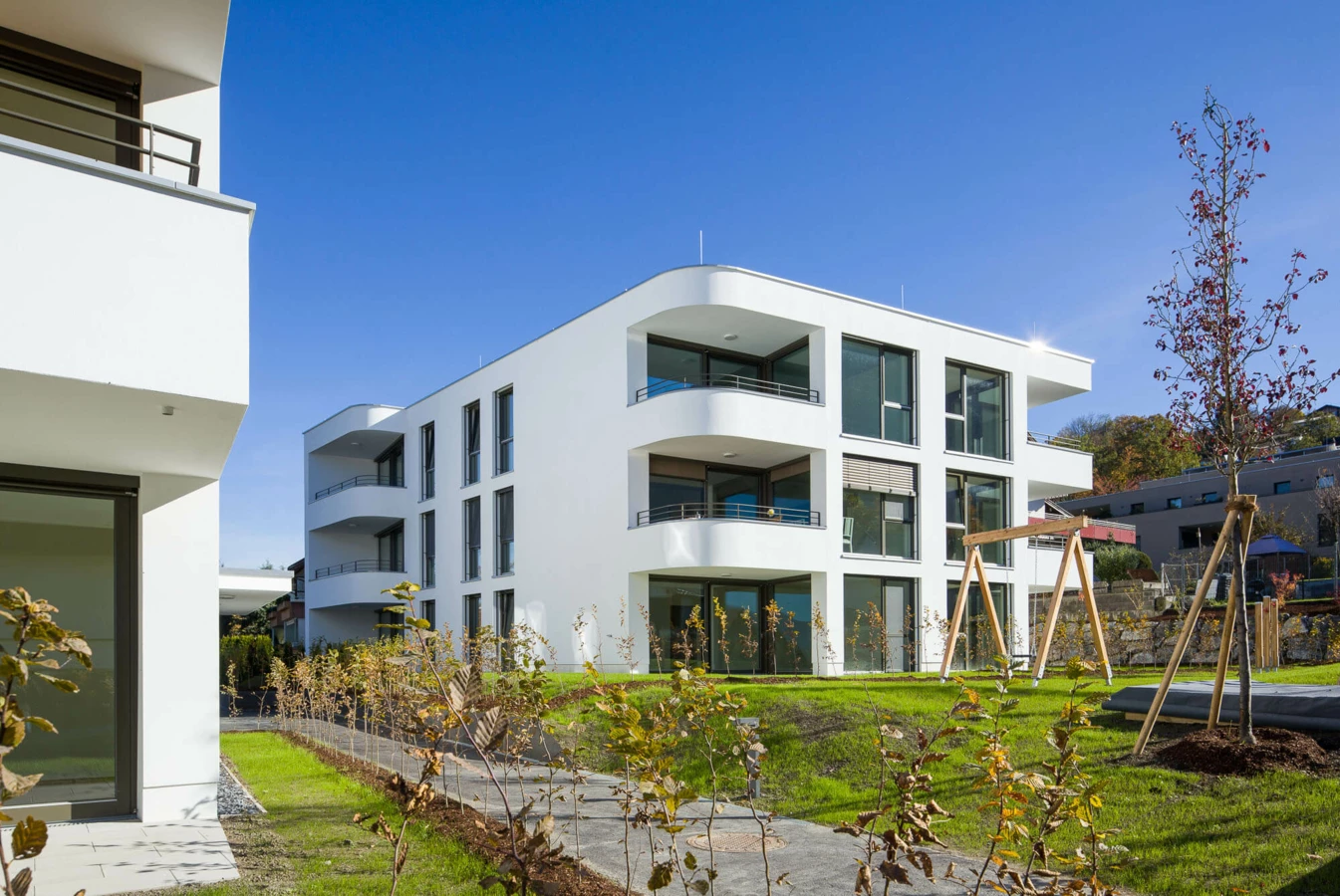 Moderne weiße Wohnanlage in Feldkirch mit gerundeten Balkonen, großen Fenstern, gepflegtem Rasen und einem Holzschaukelgerüst unter blauem Himmel (Wolf-Huber-Straße).